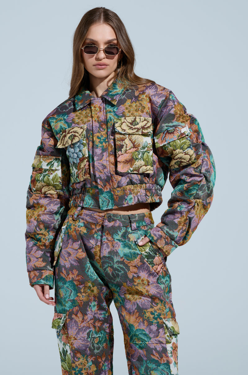 Someone confidently poses in a LOGAN SQUARE JACQUARD BOMBER, featuring colorful floral patterns and large pockets, reminiscent of tapestry. They have long hair and sunglasses against a soft blue background.