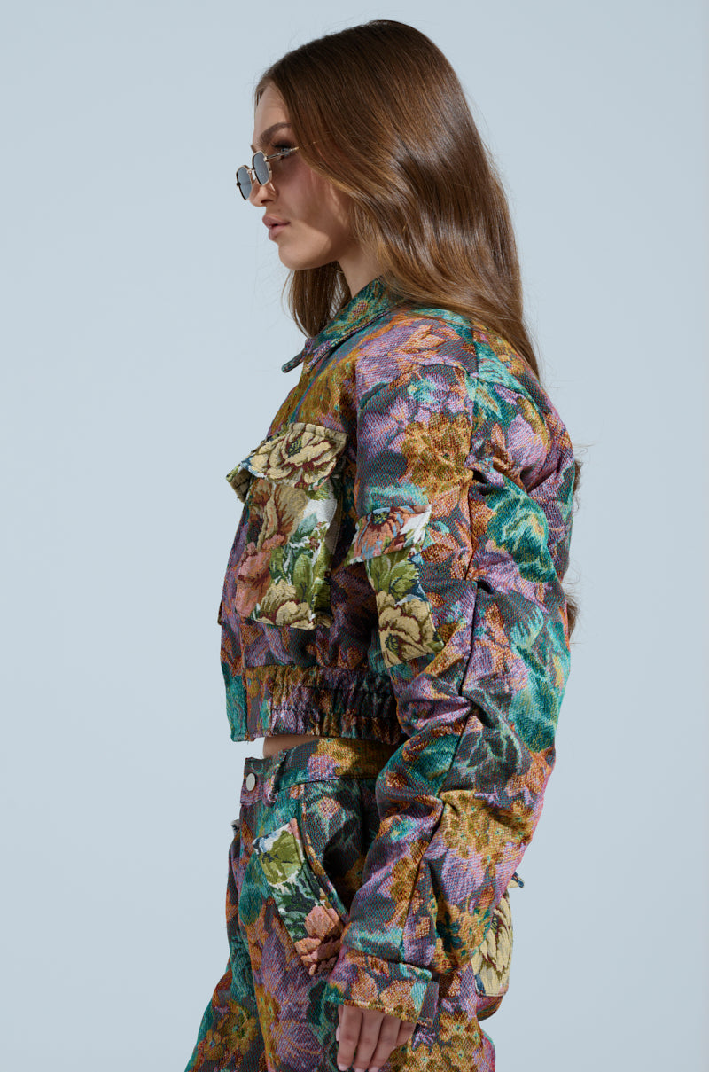 The individual with long brown hair and round glasses dons the LOGAN SQUARE JACQUARD BOMBER, showcasing a tapestry-like material in a vibrant floral pattern of purple, green, and orange hues. The ensemble includes matching pants with subtle ruched detailing against a plain backdrop.