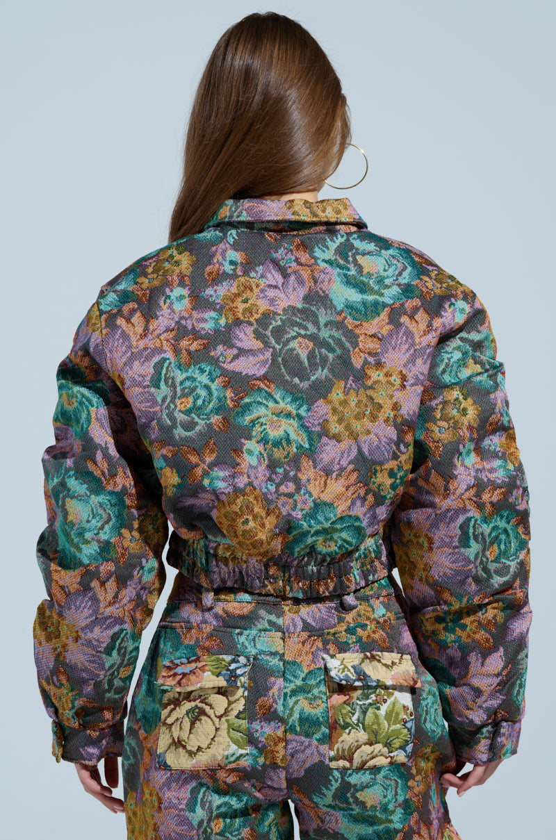 A person with long brown hair wears the LOGAN SQUARE JACQUARD BOMBER with matching pants, displaying floral-patterned, tapestry-like material vibrant in green, purple, and orange hues. The outfit features ruched detailing as they stand facing away against a plain blue background.
