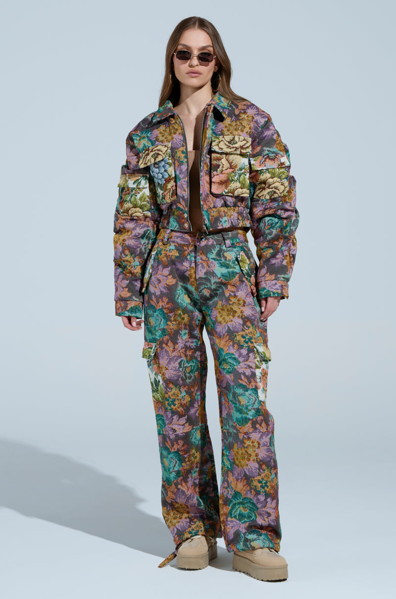 Wearing the LOGAN SQUARE JACQUARD BOMBER, a person in a vivid floral jacket and pants with tapestry-style fabric stands confidently against a light blue background. They sport long hair, sunglasses, and finish the look with beige platform shoes.