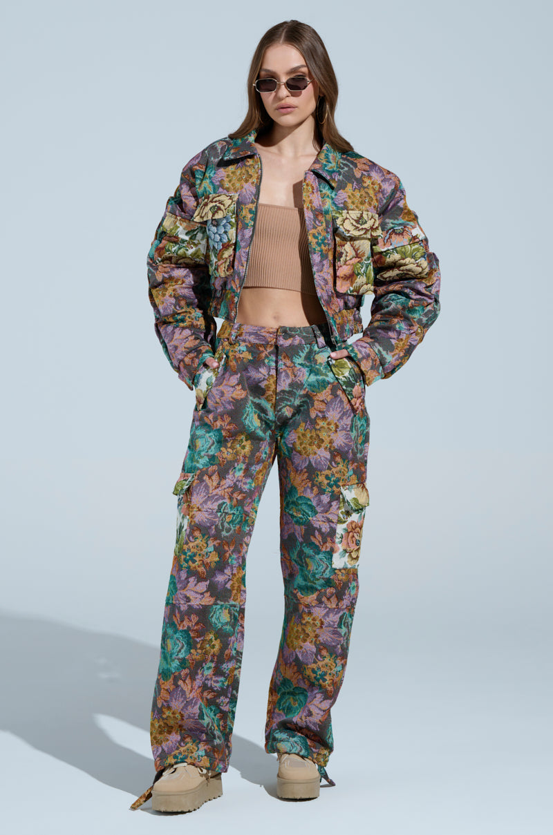 Dressed in a Logan Square Jacquard Bomber with ruched detailing, floral cargo pants, and beige platform shoes, a confident individual stands against a light blue background. With sunglasses and hands in pockets, they exude effortless style.