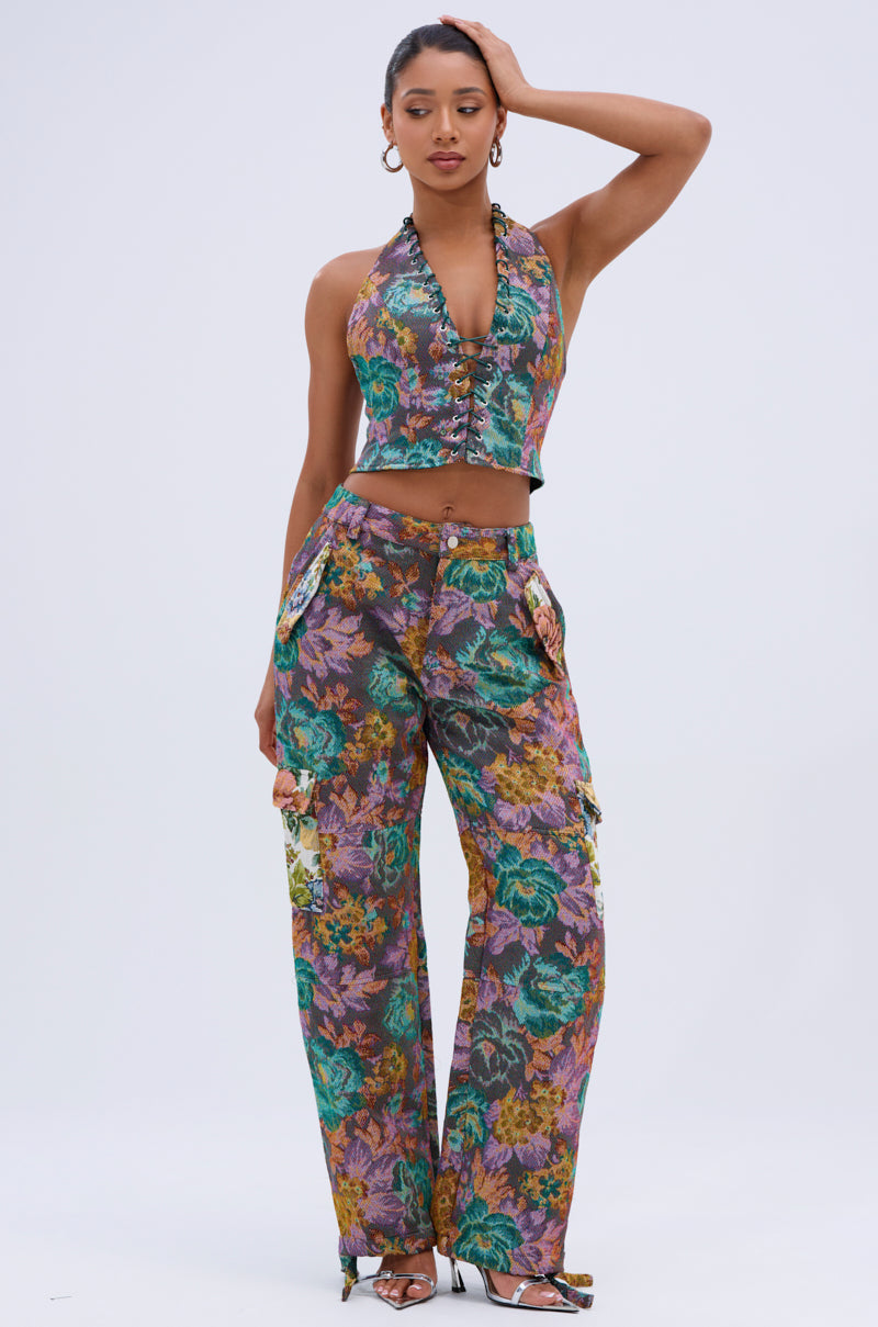 A woman poses against a plain background in an AKIRA Label sleeveless floral halter top and matching LOGAN SQUARE JACQUARD CARGO PANT with side pockets. She rests one hand on her head and pairs the look with open-toe heels.