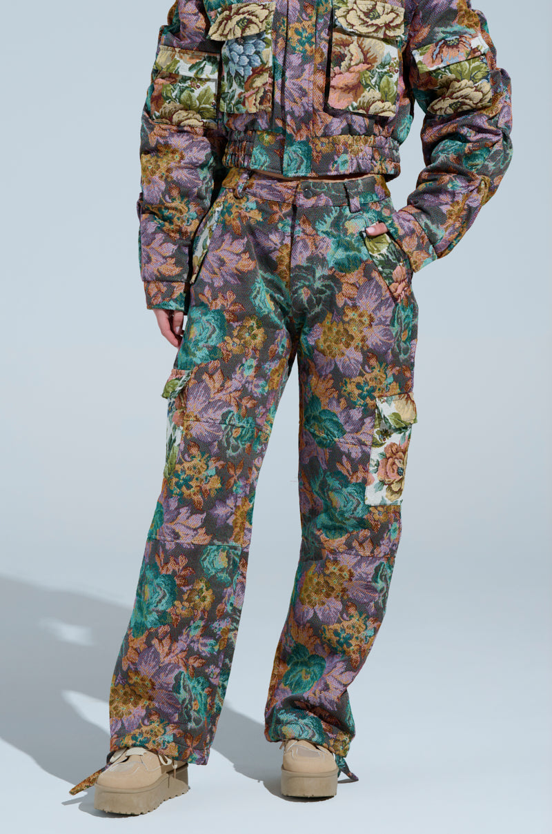 A model displays the colorful LOGAN SQUARE JACQUARD CARGO PANT ensemble, featuring a floral-patterned jacket and wide-legged cargo pants with pockets in green, purple, and brown hues. They rest their hands in the pockets while wearing tan shoes.