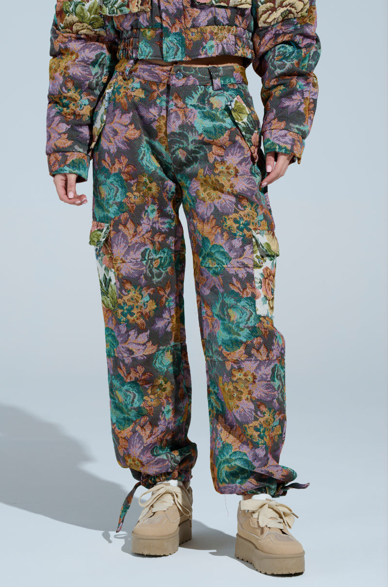 Clad in matching purple, green, and brown floral-patterned attire, a person wears a jacket and the Logan Square Jacquard Cargo Pant. Made from an upholstery-like fabric, they pose against a light blue backdrop, finishing the look with beige boots.