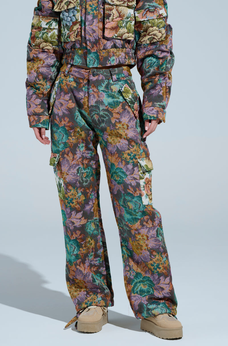 Styled in a camo-patterned puffer jacket with matching LOGAN SQUARE JACQUARD CARGO PANT, a person stands against a light blue background in tan boots. The jacket's upholstery-like material and multiple pockets enhance its functional and aesthetic appeal, featuring green, brown, and purple tones.