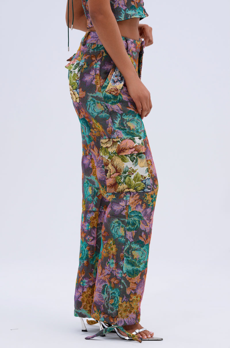 A person stands sideways in heels against a plain light background, wearing the LOGAN SQUARE JACQUARD CARGO PANT with a colorful floral tapestry pattern and matching crop top, both featuring elasticized strap closures.