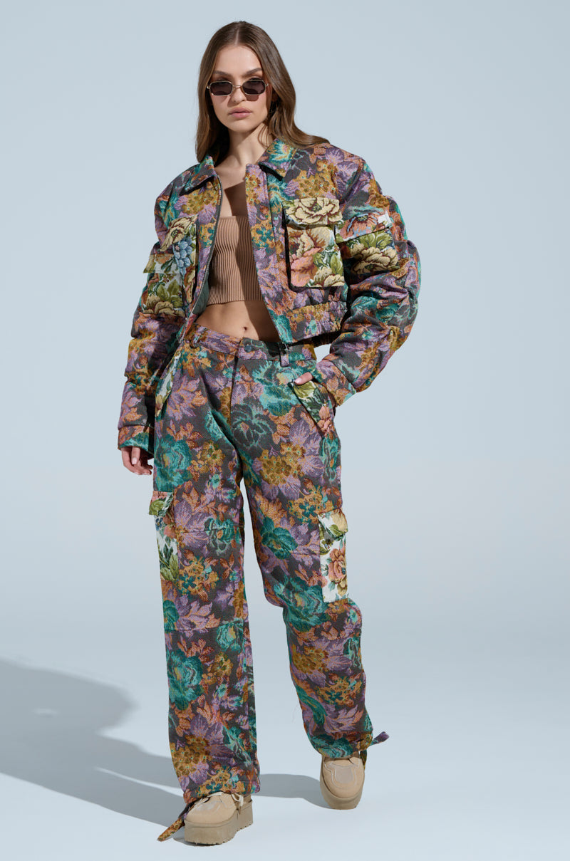 Wearing sunglasses, a person confidently poses in a Logan Square Jacquard floral outfit featuring matching jacket and cargo pants from the AKIRA Label in green, purple, and brown. A ribbed beige top and beige shoes complete the look.
