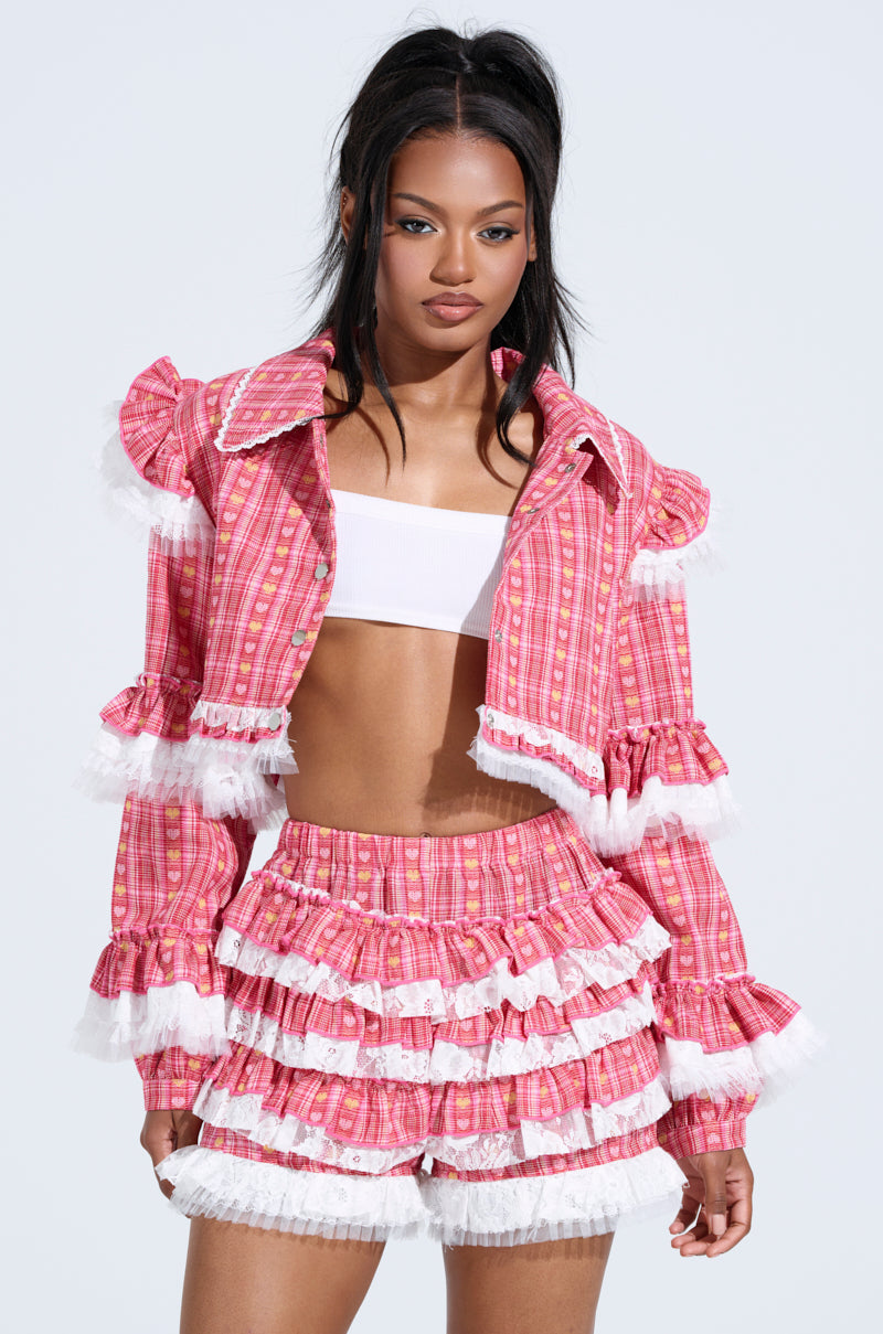 Against a light blue background, a woman poses confidently in the SUNNY RUFFLE BOMBER—a pink plaid set with ruffled details and lace trim, featuring a cropped jacket and matching shorts. Her long dark hair is styled in a high ponytail.