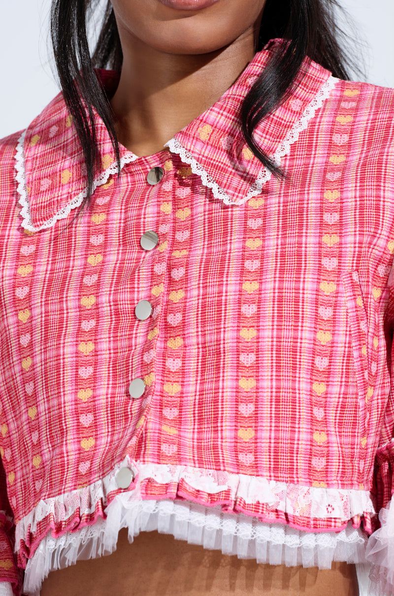 Close-up of someone wearing a red plaid cropped shirt with heart patterns and lace trim, styled like the SUNNY RUFFLE BOMBER. It features a large collar, snap buttons down the front, and showcases intricate details, with the person's face partially visible.