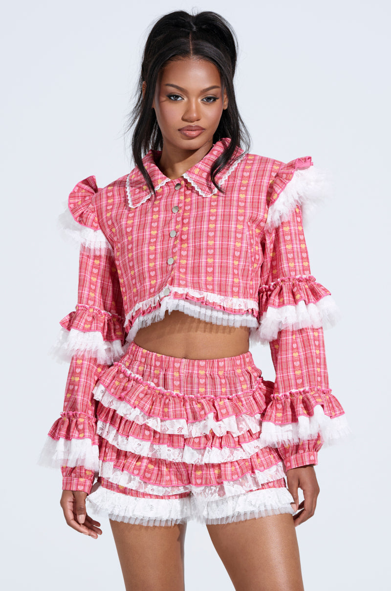 A woman wears the SUNNY RUFFLE BOMBER, a pink ruffled ensemble with a plaid pattern and white lace trim. Featuring a long-sleeve cropped top and matching high-waisted shorts, it creates a vibrant, chic look against the plain backdrop.