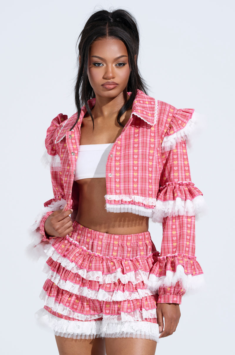 A woman models the Sunny Ruffle Bomber, featuring a pink/white plaid pattern with lace trim, ruffled shoulders and sleeves. Her stylish outfit includes a cropped jacket and shorts ensemble as she poses elegantly against a plain background.