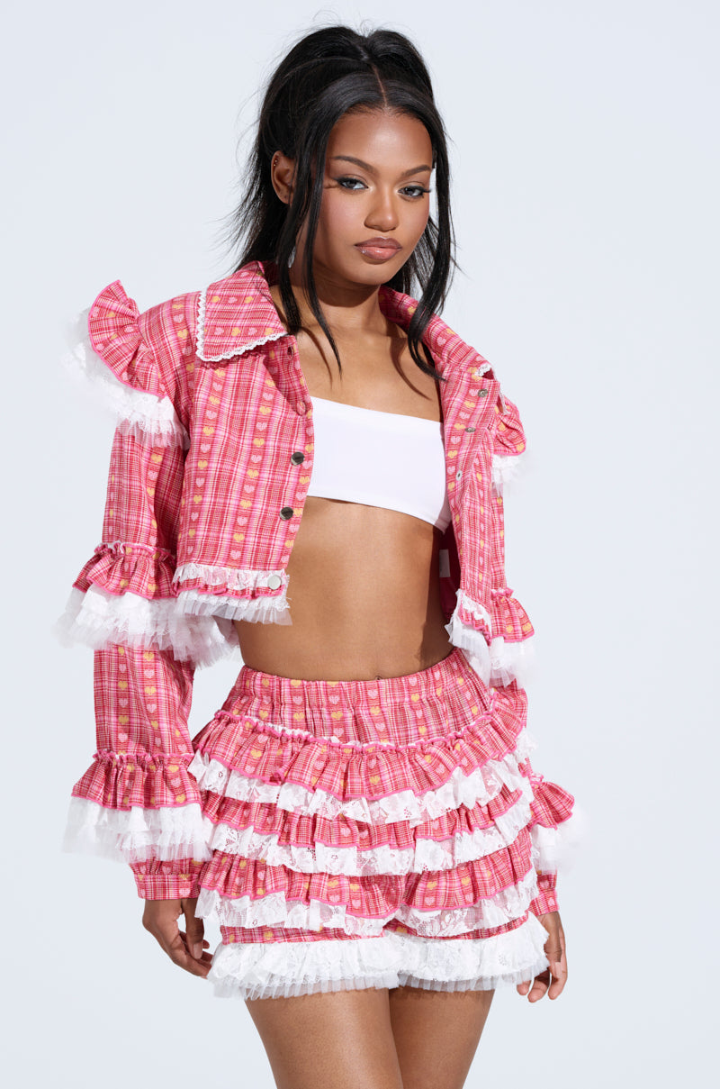 A woman stuns in a pink plaid ensemble with a crop top and the Sunny Ruffle Bomber jacket, paired with a ruffled skirt. White lace trim adds elegance, while her long dark hair is styled chicly half-up, half-down, set against a plain backdrop.