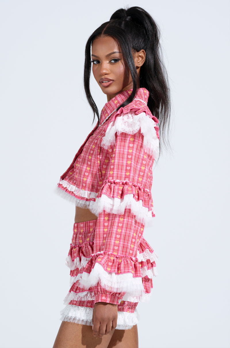 A woman in the SUNNY RUFFLE BOMBER, a pink plaid with white frills, stands in side profile. Her half-up hairdo is accented with a delicate lace trim as she gazes confidently at the camera against a plain background.
