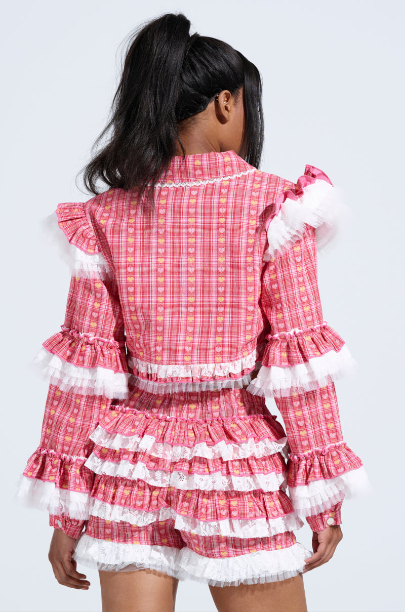 A person with long, dark hair in a ponytail wears the SUNNY RUFFLE BOMBER, a pink plaid outfit featuring ruffles and delicate lace trim. Facing away from the camera, they highlight the layered, textured design on the sleeves and shorts.