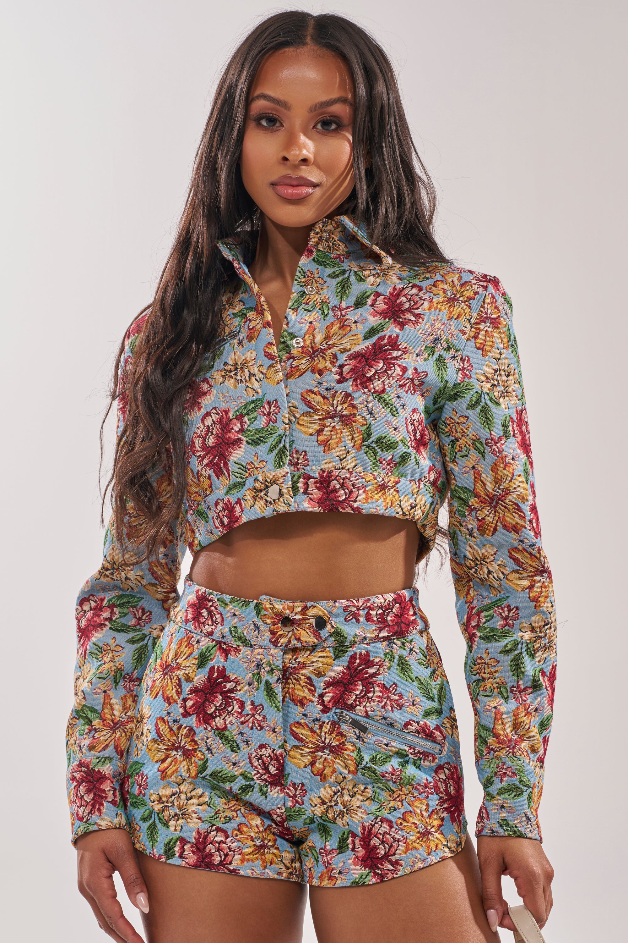 A woman with long dark hair wears the GOOD KARMA TAPESTRY MOTO IN BLUE MULTI, a matching floral outfit with a cropped, long-sleeved button-up top and high-waisted shorts adorned with red, yellow, and white flowers on blue. 0