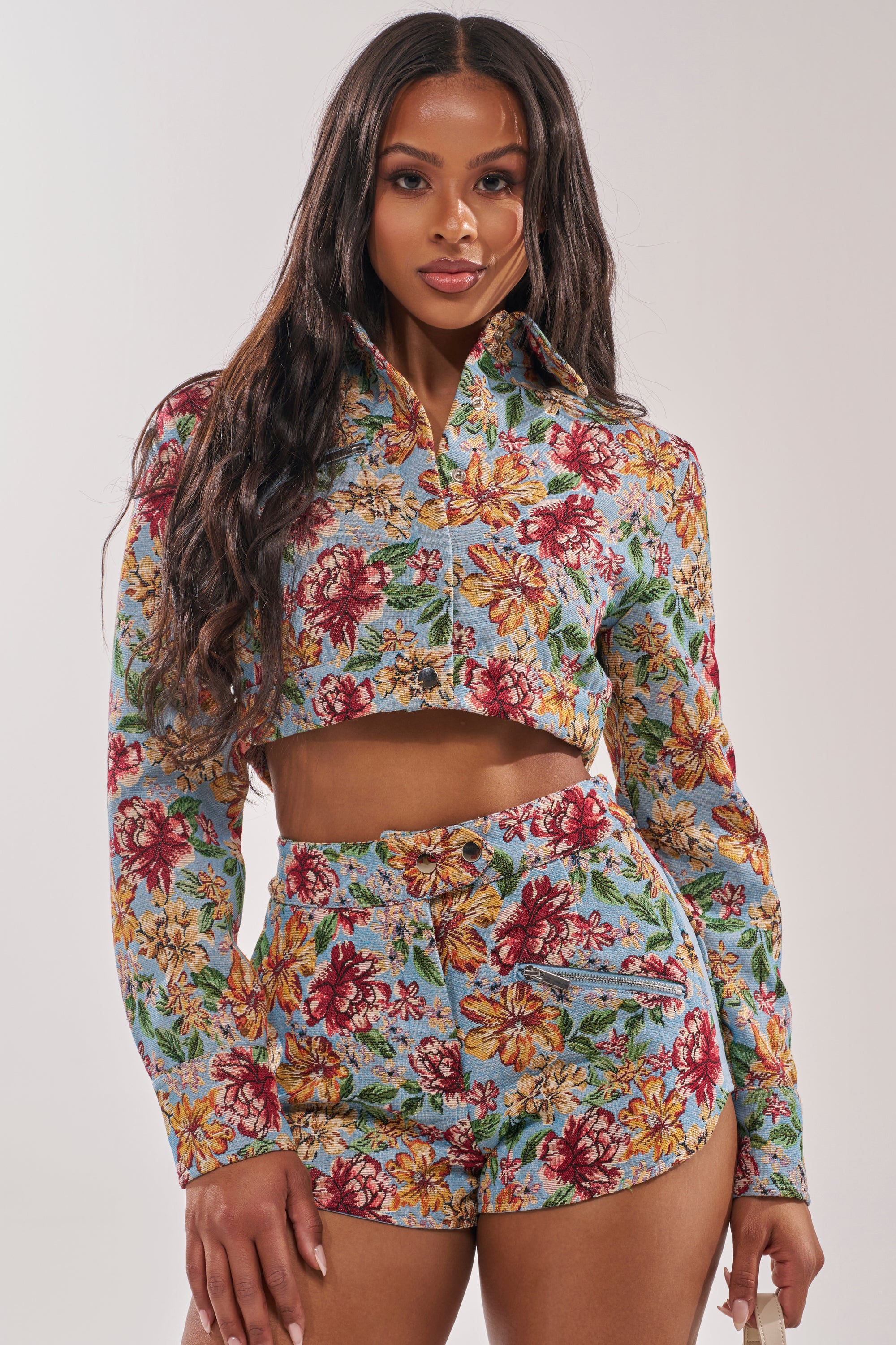 A woman stands confidently against a plain light background, wearing the GOOD KARMA TAPESTRY MOTO IN BLUE MULTI—a cropped floral tapestry jacket with an ultra-cropped hem and matching high-waisted shorts in blue, red, yellow, and green. 2