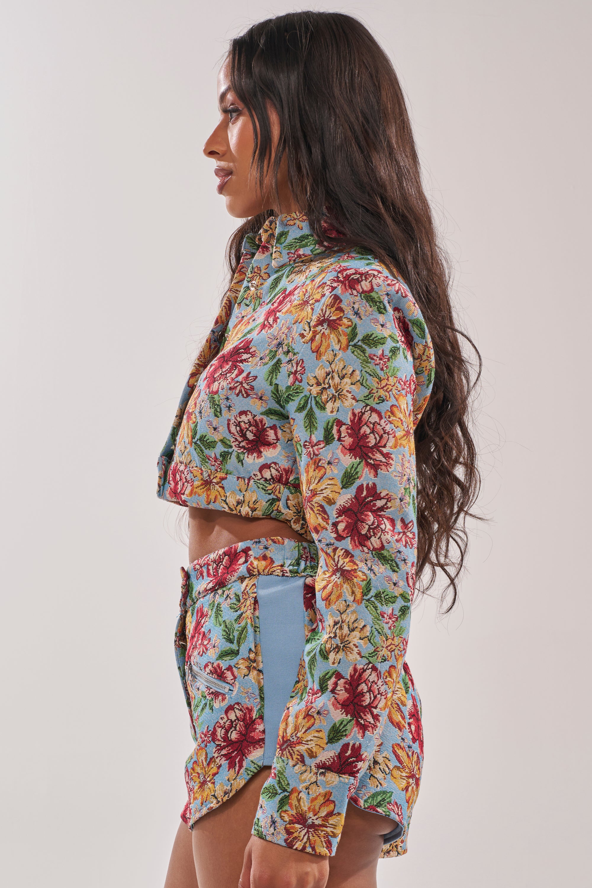 A woman with long wavy brown hair stands sideways, wearing the GOOD KARMA TAPESTRY MOTO IN BLUE MULTI jacket and matching high-waisted shorts. She poses against a plain, light-colored background. 3