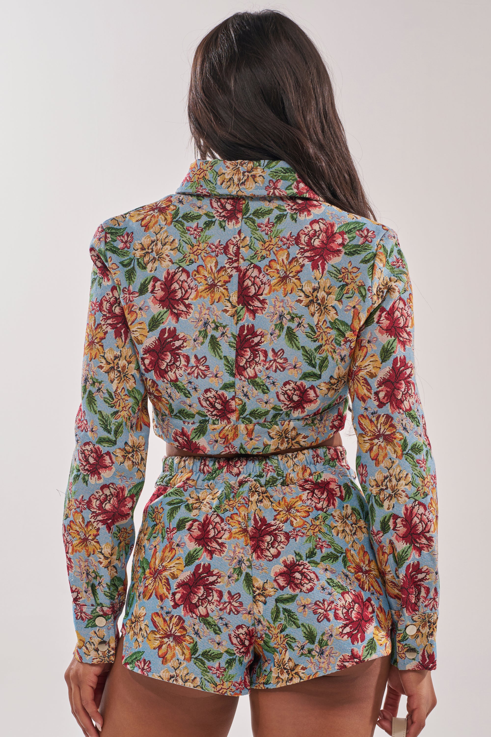 A woman with long dark hair is shown from behind, wearing the GOOD KARMA TAPESTRY MOTO IN BLUE MULTI— a cropped floral jacket with satin lining— over a matching colorful floral shirt and shorts set. 4