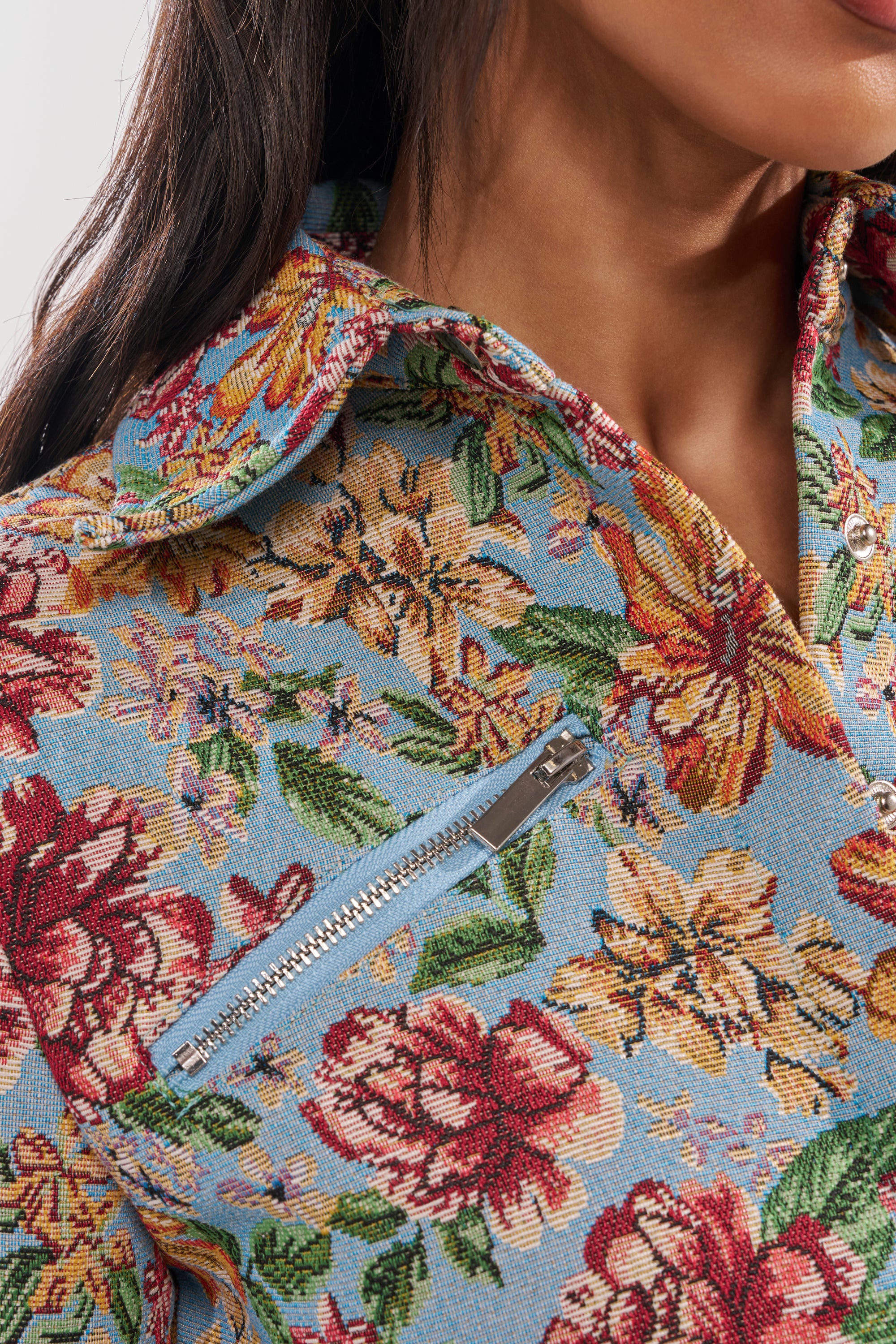 Close-up of a woman wearing the GOOD KARMA TAPESTRY MOTO IN BLUE MULTI, featuring a cropped floral tapestry design, ultra-cropped hem, and diagonal zip chest pocket. Her long dark hair is partially visible as she smiles softly. 7