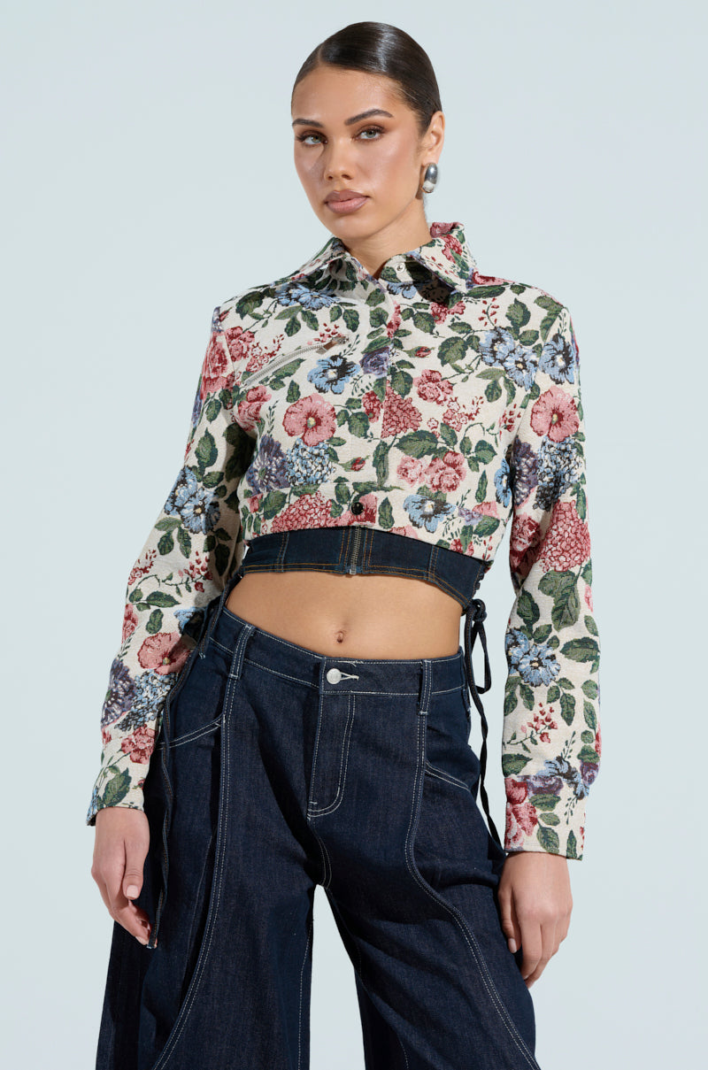 A person poses confidently in the GOOD KARMA TAPESTRY MOTO floral print crop top with a collar and high-waisted dark jeans, highlighting a chic cropped look. The background is a plain light color, and their hair is styled neatly.