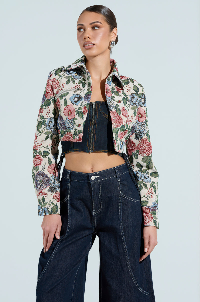 A person poses confidently in the GOOD KARMA TAPESTRY MOTO jacket over a black top and dark wide-legged jeans against a light background, with hair pulled back and gaze directed slightly to the side.