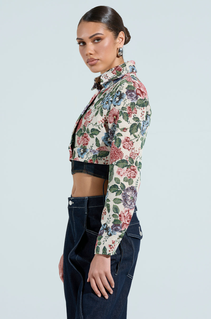 Facing left against a light background, the person showcases a floral Good Karma Tapestry Moto jacket with dark blue jeans. Their ponytail adds elegance to the vibrant look.
