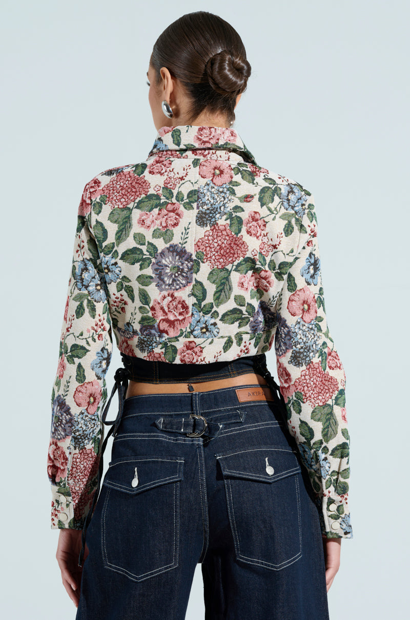 A person with a neat bun wears the Good Karma Tapestry Moto jacket adorned with red, pink, and blue flowers. They pair it with dark high-waisted jeans, highlighting their chic cropped look against a light blue backdrop.