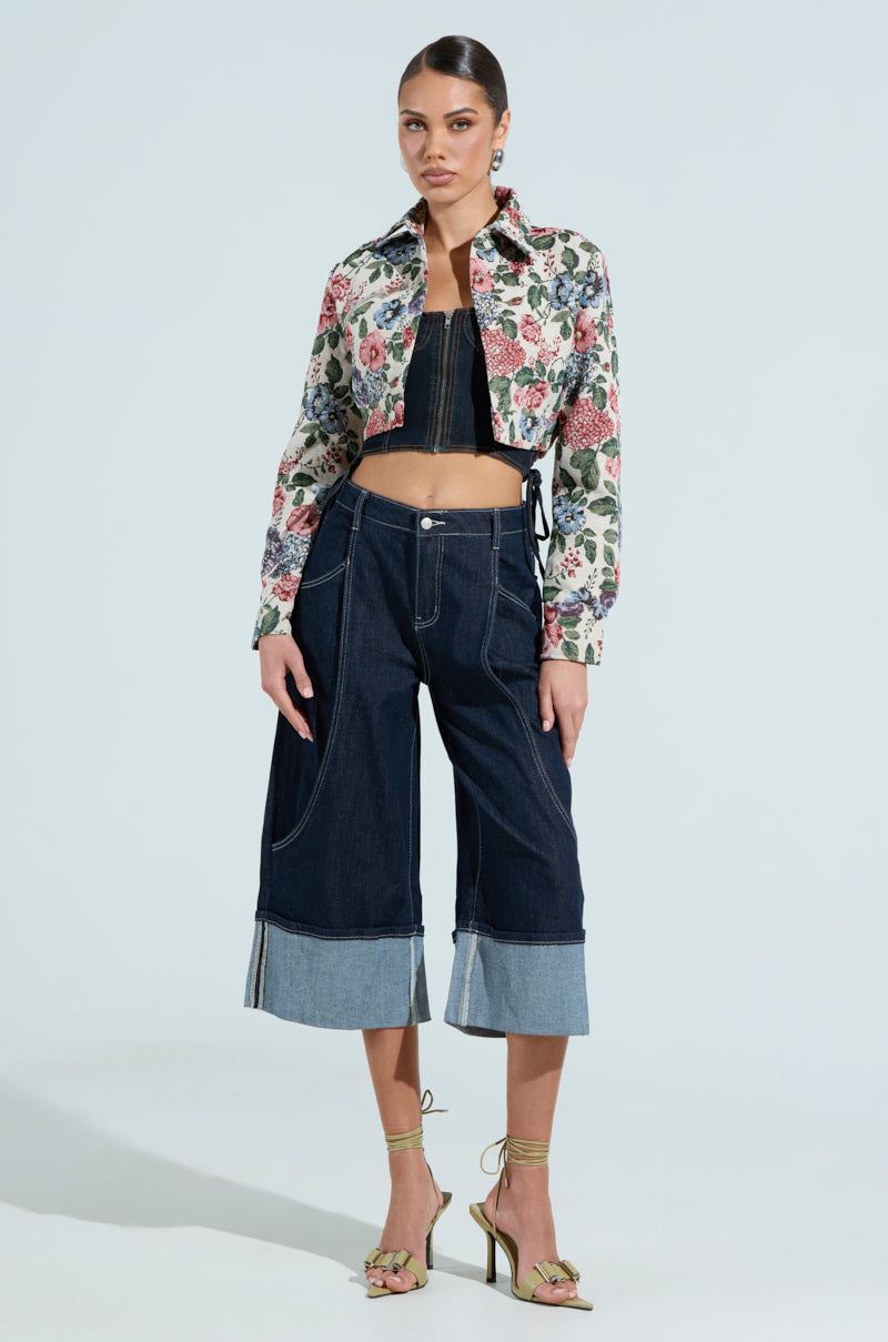 A person models a floral cropped silhouette jacket over a black top with wide-leg denim jeans, paired with green strappy heels and sleek pulled-back hair. The light blue background perfectly complements their Good Karma Tapestry Moto look.