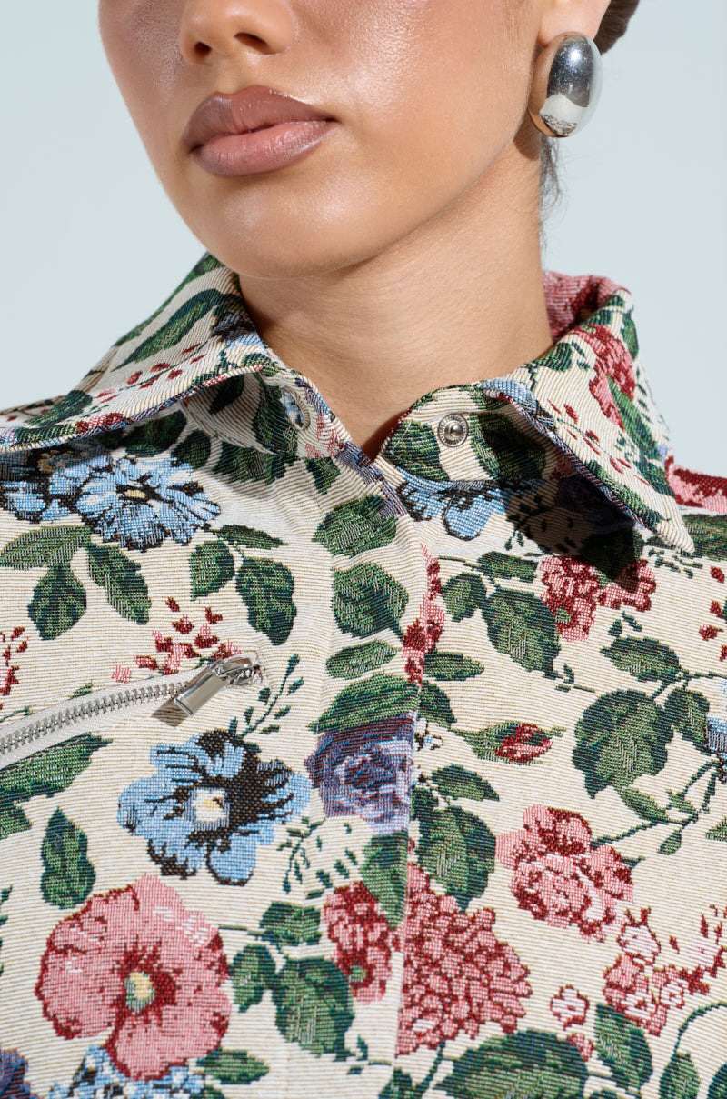 A close-up of a person wearing the GOOD KARMA TAPESTRY MOTO jacket, featuring vibrant red, blue, and green floral prints on a cream background with shiny buttons. They also wear silver hoop earrings. Their lips are closed as they slightly turn their head, showcasing the cropped silhouette.
