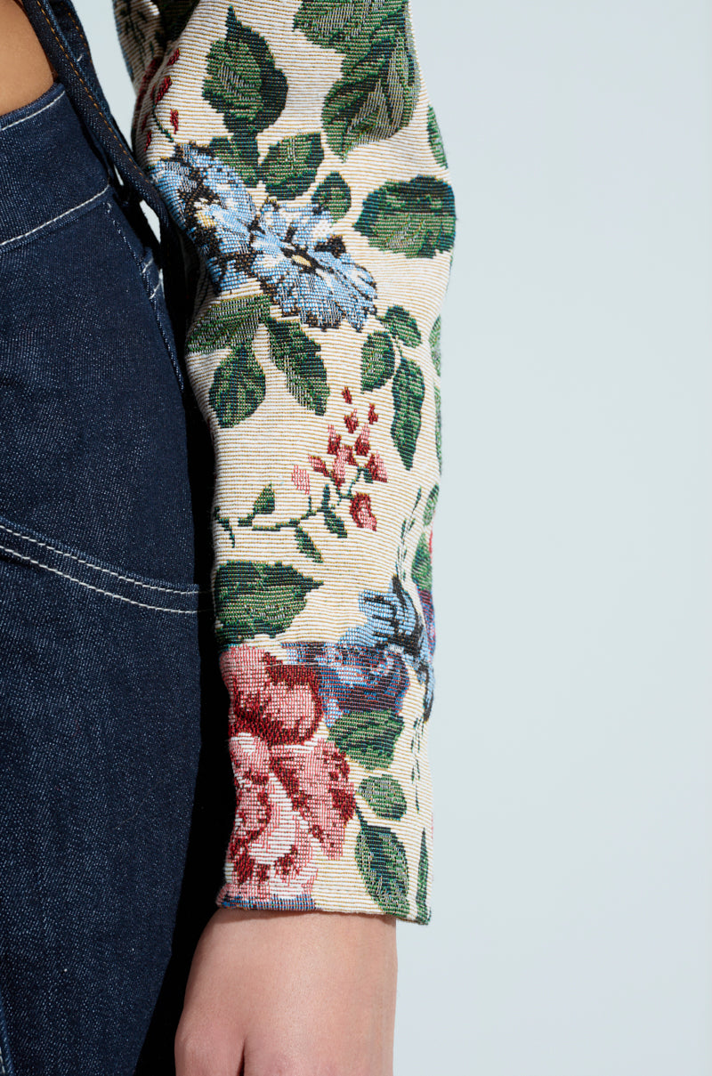 A close-up of someone in dark blue jeans and a floral top similar to the Good Karma Tapestry Moto, featuring vibrant red, blue, and green flowers on a light background, with one hand resting by their side.