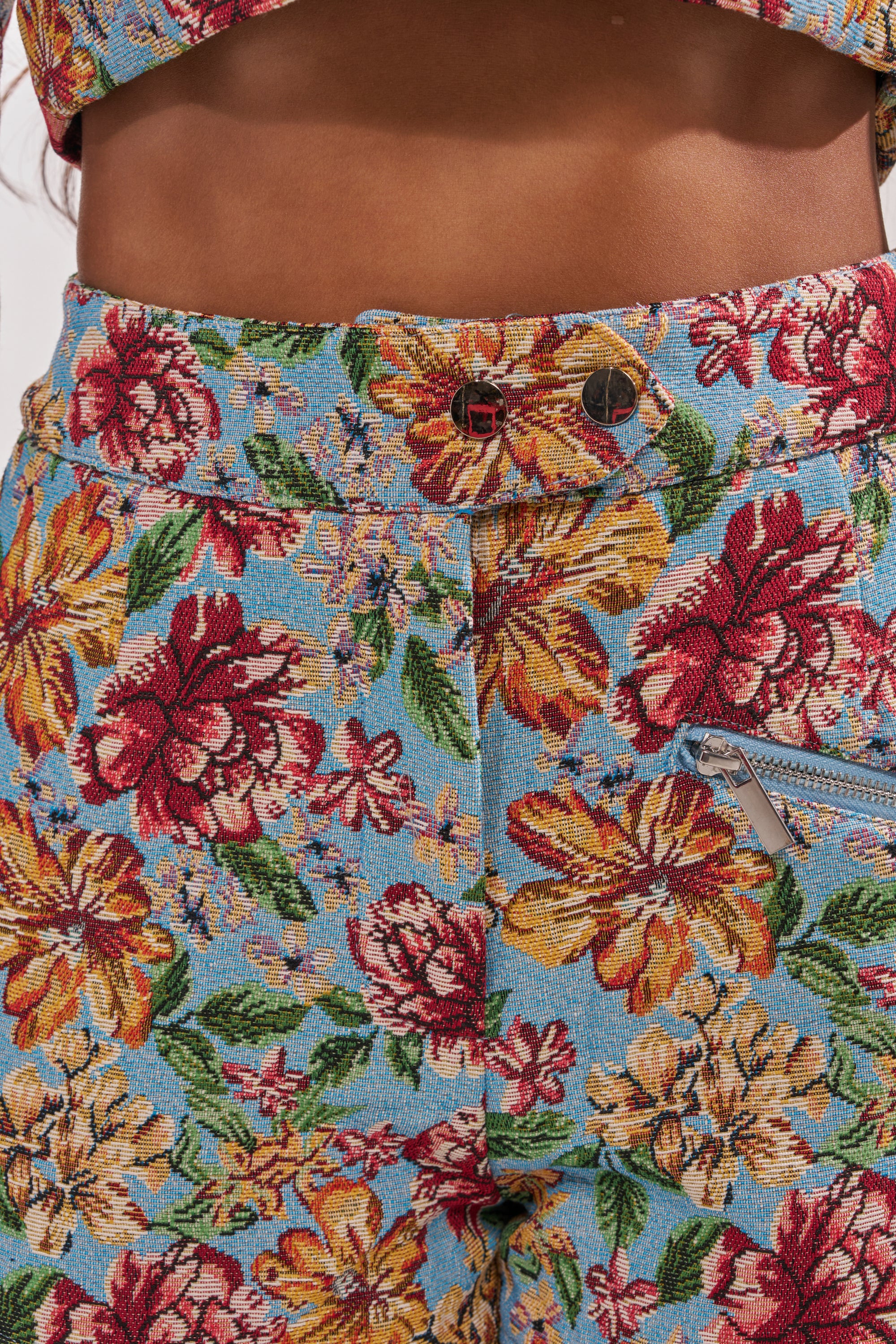 Close-up of a person wearing the GOOD KARMA TAPESTRY SHORTS IN BLUE MULTI, featuring a high-rise waist, bold floral pattern in red, yellow, and orange, front zipper, silver side pocket zipper, and double button closure for vintage flair. 7