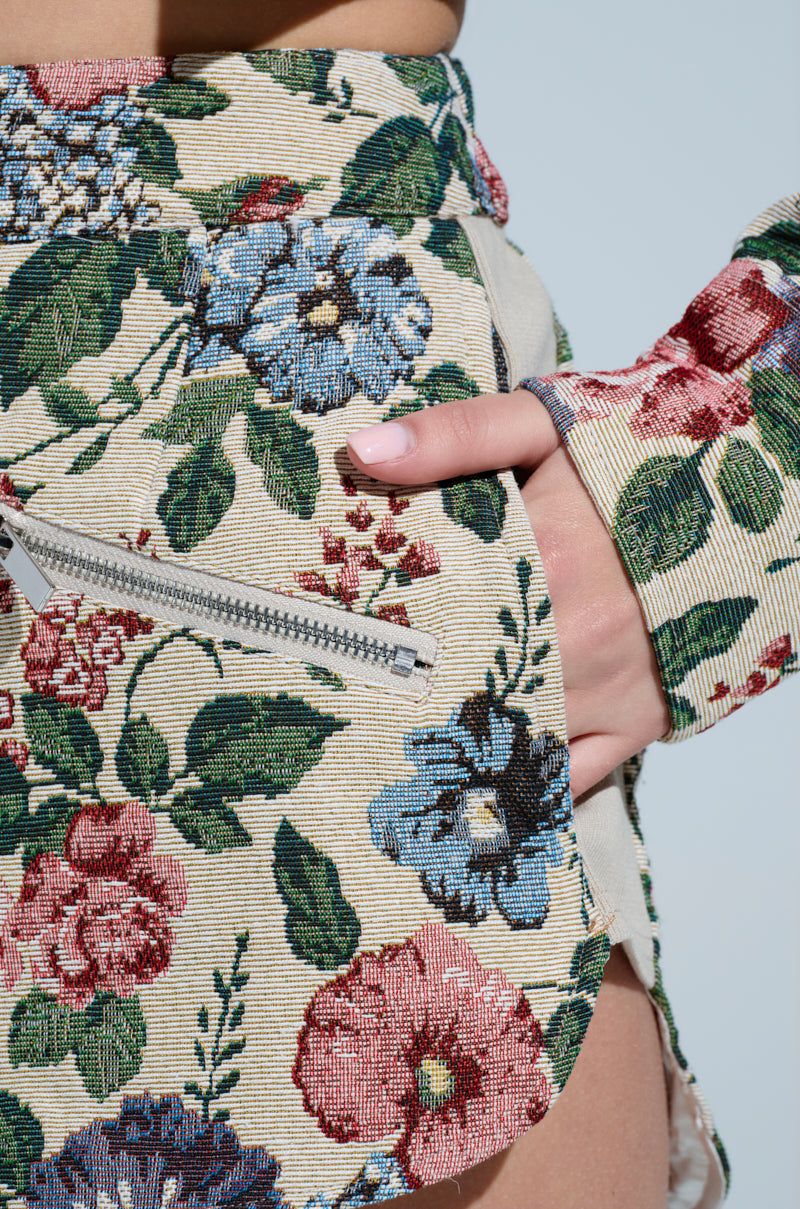 A close-up features someone in high-rise GOOD KARMA TAPESTRY SHORTS with a vibrant floral design of red, blue, and green flowers. A hand touches a pocket with a visible side zipper against a soothing light blue background.