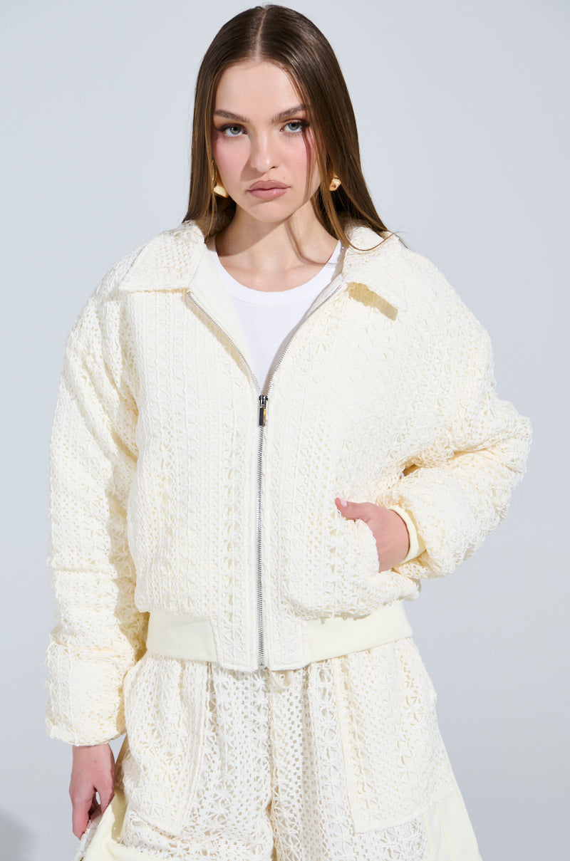 A woman with long brown hair models the AKIRA Label's BROOKLYN CROCHET BOMBER, which has a quilted inner lining. She stands against a plain, light background, one hand in her pocket, gazing at the camera.