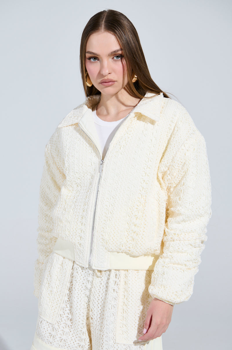 Dressed in a cream-colored textured jacket similar to the BROOKLYN CROCHET BOMBER and matching pants, a person with long hair showcases their chic style. A white top peeks beneath the jacket against a plain, light background that enhances their effortlessly stylish appearance.