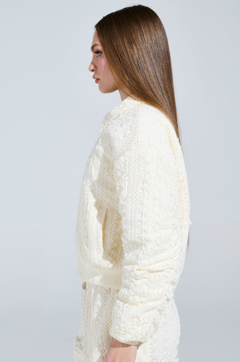A person with long, straight hair stands in profile against a plain background, donning the BROOKLYN CROCHET BOMBER by AKIRA Label—a textured, cream-colored outfit comprising a collared top with long sleeves and quilted inner lining. The look is sleek and modern.