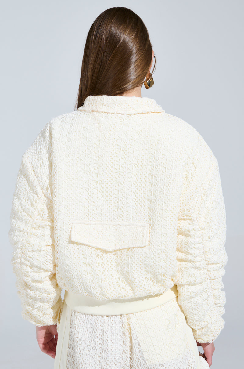 The image shows a person with long brown hair viewed from behind, wearing an off-white textured coat and matching skirt. The coat, similar to the BROOKLYN CROCHET BOMBER style, has a rectangular back flap while the person casually holds one hand at their side.
