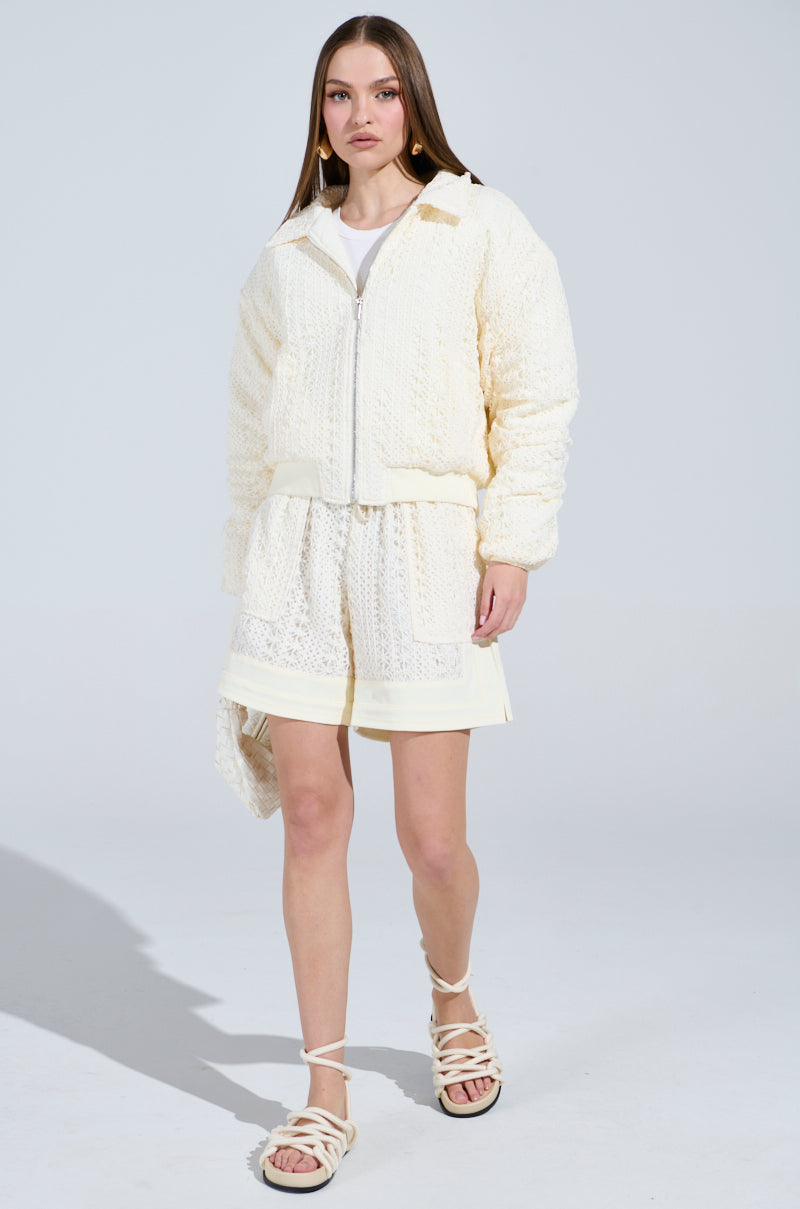 A woman models the BROOKLYN CROCHET BOMBER, a cream-textured jacket with a quilted lining, paired with matching shorts and a white top. White sandals complete the look against a plain light gray background.