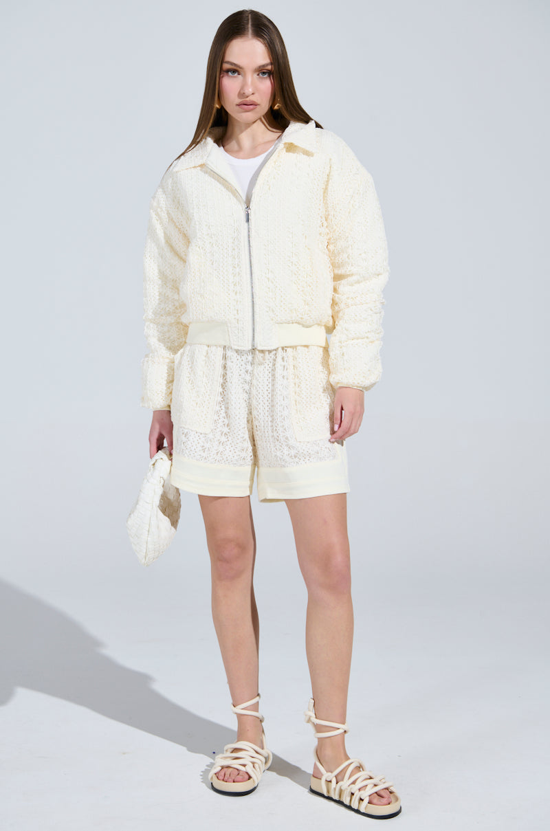 Wearing the AKIRA Label BROOKLYN CROCHET BOMBER and matching shorts over a white top, with a small matching bag and cream sandals, the person stands against a plain light-colored background. The bomber's quilted inner lining adds texture.