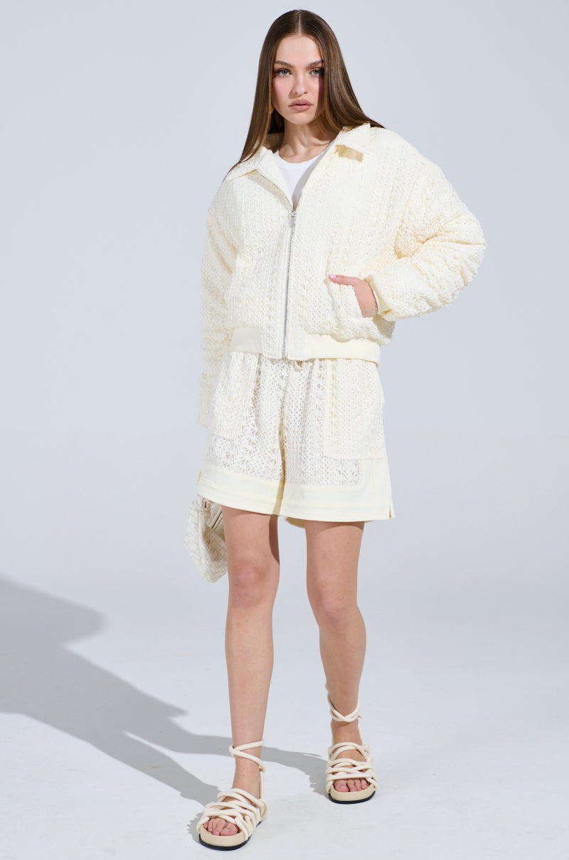 Wearing the BROOKLYN CROCHET BOMBER, a cream-textured jacket with quilted lining by the AKIRA Label, they pair it confidently with matching shorts and sandals. Posing against a plain backdrop, they gaze directly at the camera.