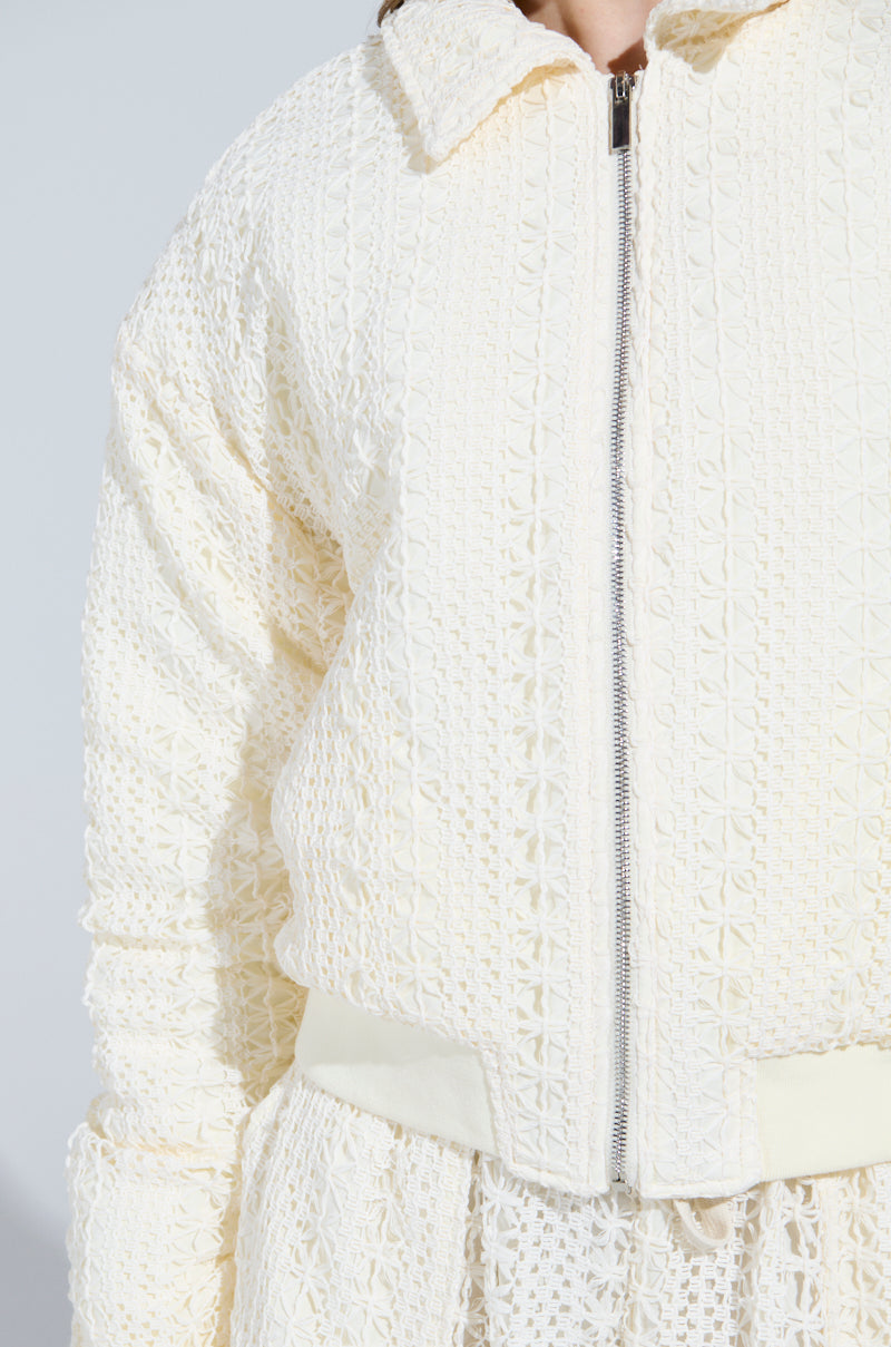 A close-up shows a person wearing the textured cream AKIRA Label BROOKLYN CROCHET BOMBER, featuring intricate patterns and a front zipper, paired with a matching skirt. The quilted inner lining enhances comfort against a plain, light background.