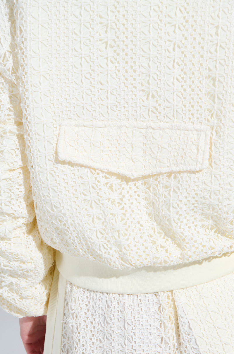 A person is seen from the back wearing an AKIRA Label outfit in cream, knitted with ribbed edges and a rectangular pocket. The stylish BROOKLYN CROCHET BOMBER design resembles a skirt with a waist belt, adding flair.