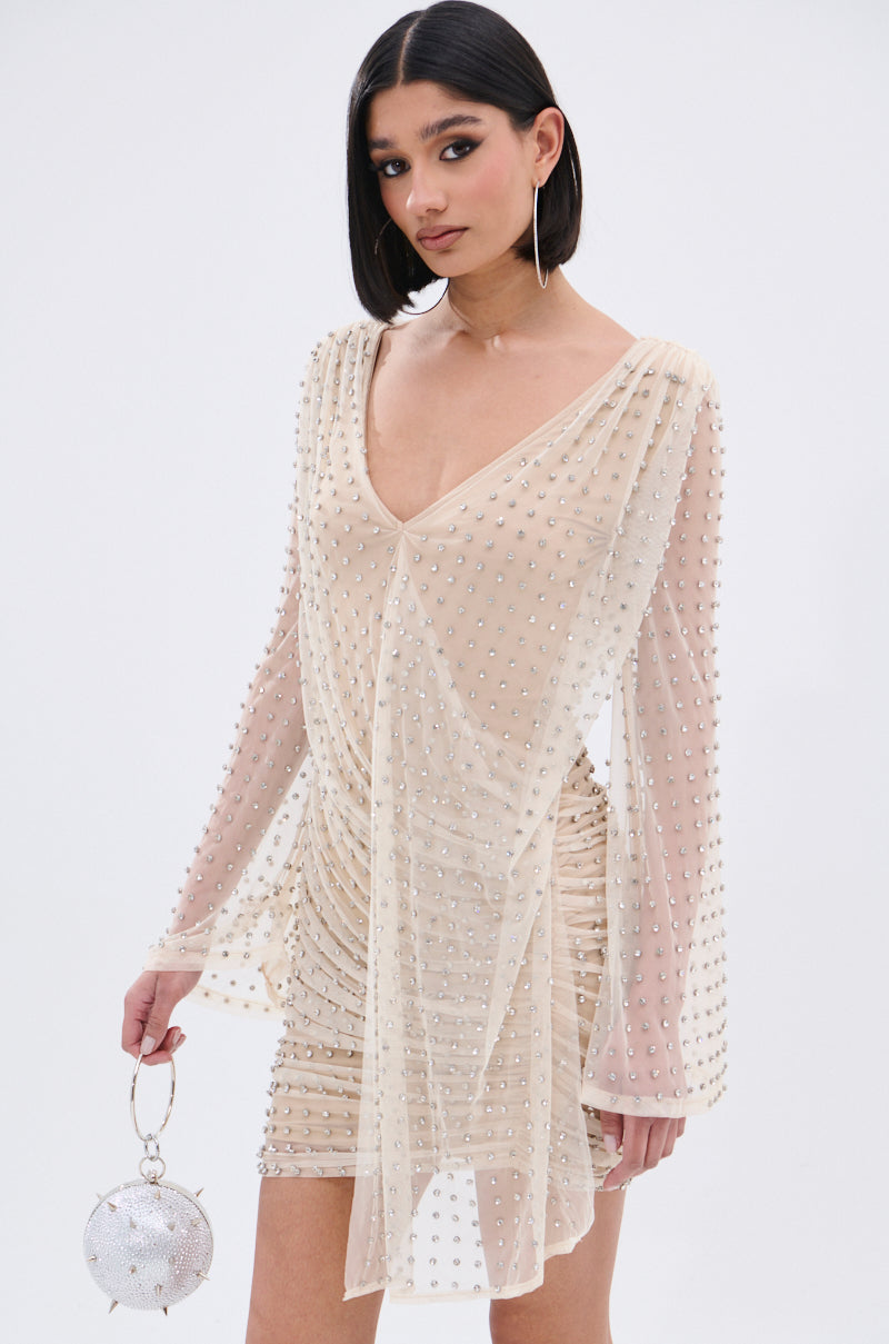 A woman stands against a white background wearing the ALYSSA RHINESTONE MINI DRESS, featuring nude mesh, ruching, and rhinestone accents. She has a sleek bob, large hoop earrings, and carries a round silver handbag.