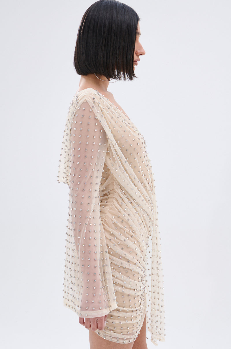 Woman with a sleek black bob in the ALYSSA RHINESTONE MINI DRESS, a sheer nude mesh long-sleeve style adorned with sparkling embellishments, stands in profile against a plain light background.