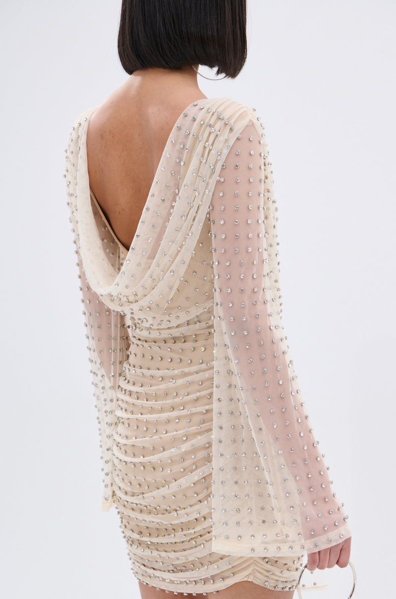 A woman with shoulder-length dark hair wears the ALYSSA RHINESTONE MINI DRESS, a beige nude mesh mini with long sleeves, pearl embellishments, a ruched design, and a low draped back that elegantly reveals her back and arms.