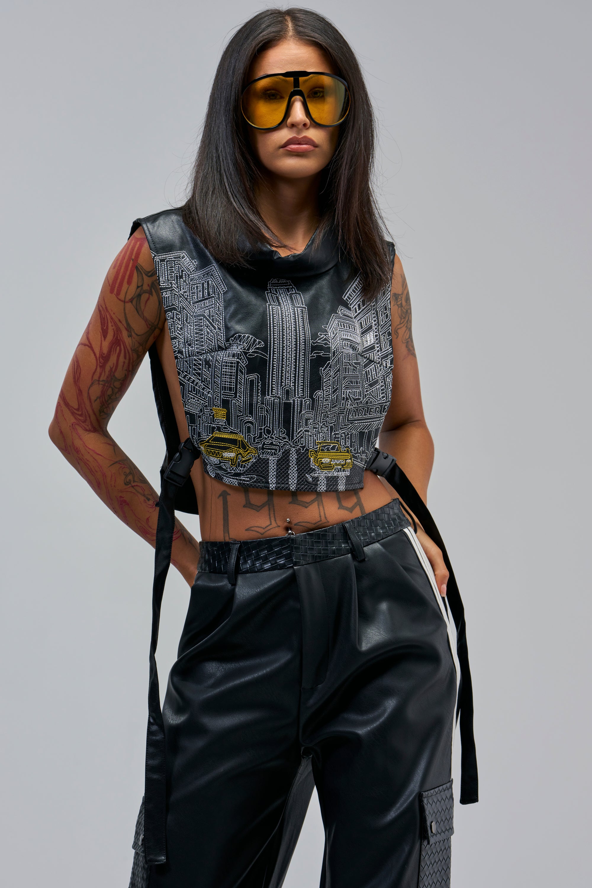 A woman with long dark hair, yellow sunglasses, and tattoos rocks festival style in the CITY STREETS PU TOP and black leather pants, standing confidently against a plain gray background.