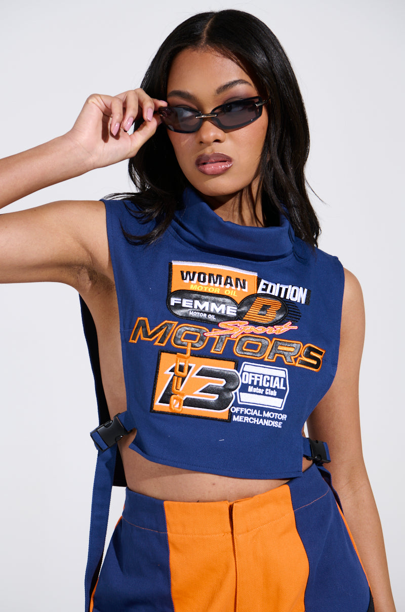 Sporting the MOTORS TOP with racing-themed graphics, a person in dark sunglasses poses confidently in high-waisted, two-tone blue and orange pants. The cropped silhouette adds sleek style as they adjust their shades with confidence.