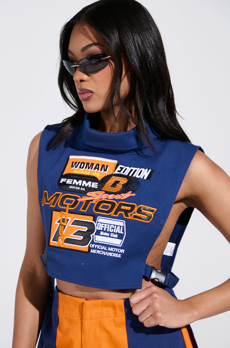 A person wears the stylish MOTORS TOP in blue and orange, featuring racing-inspired patches. With long dark hair cascading past the denim design, they confidently stand against a plain backdrop, donning sunglasses that enhance their cool, cropped silhouette.