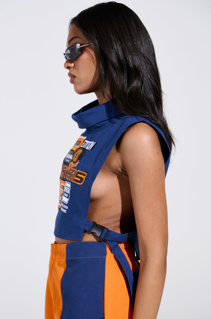 A person with long dark hair and sunglasses wears a MOTORS TOP, a blue and orange sleeveless piece adorned with racing-inspired patches and a high collar. The graphic design complements their profile against a plain white background.