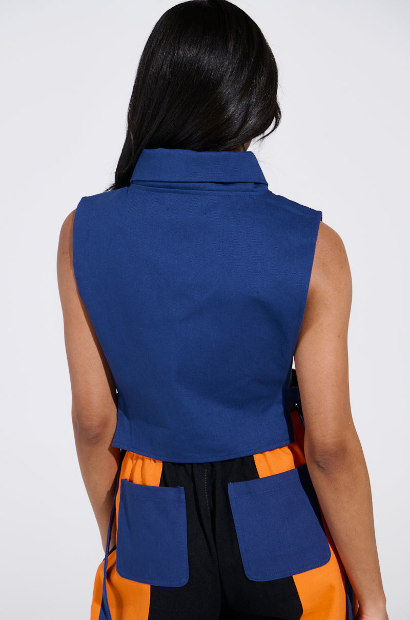 A person with long dark hair is facing away, wearing the sleeveless blue MOTORS TOP with a racing patch collar, paired with color-blocked pants in black, orange, and blue. The pants feature two blue square pockets on the back.