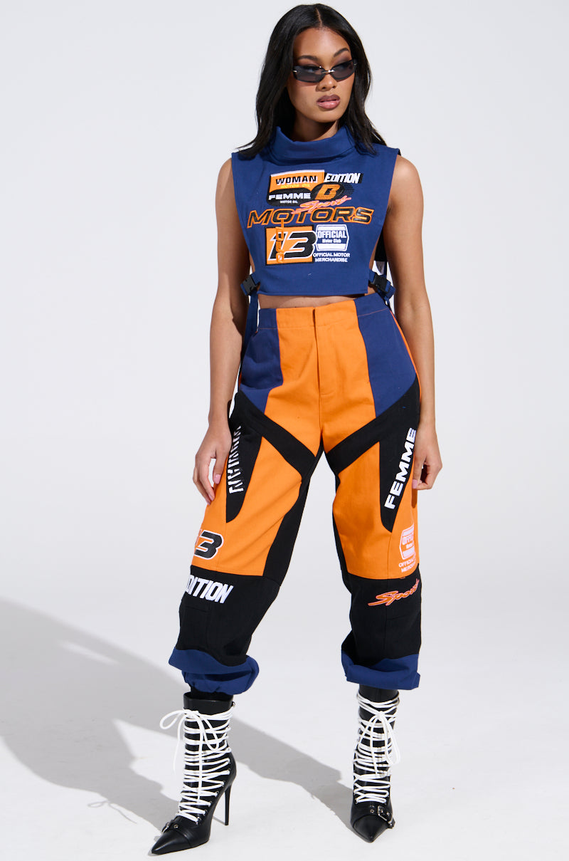 Wearing the chic MOTORS TOP with its racing-inspired design, a woman dons this blue and orange cropped silhouette with matching pants, complete with racing patches. Her outfit is stylishly paired with sunglasses and black lace-up boots against a light background.